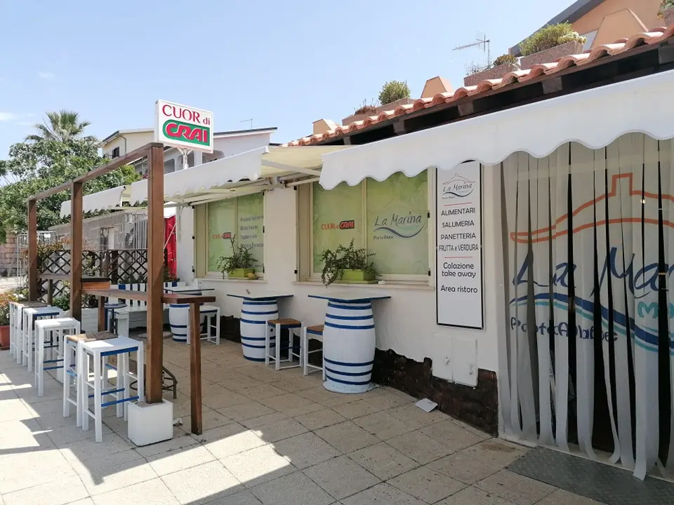 Ingresso di La Marina Market con insegna Cuor di Crai, tende bianche, botti a righe blu come tavolini e pergola in legno