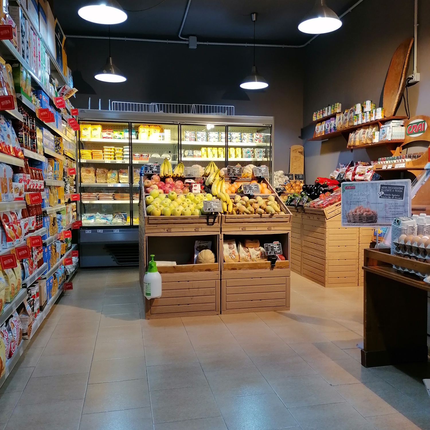 Interno di market con cassette di legno piene di frutta, frigo a vetrina, scaffali con cartelli Offerta e logo Crai
